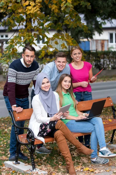 Group of young people using laptop and tablet on a park bench, having fun, talking, hanging out