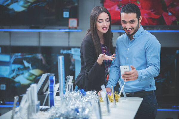 Молодая пара в магазине бытовой электроники смотрит на последние ноутбук, телевизор и фотокамеру, чтобы купить
