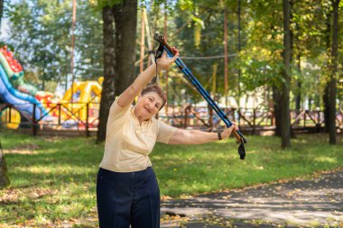 Yaşlı bir kadın, güneşli bir yaz gününde parkta sopayla kuzeye doğru yürüyor.