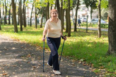 Yaşlı kadın, güneşli bir yaz gününde parkta sopayla kuzeye doğru yürüyor.