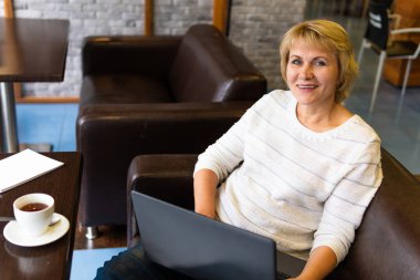 Elinde bir fincan çay ve laptopla masada oturan bir kadın.