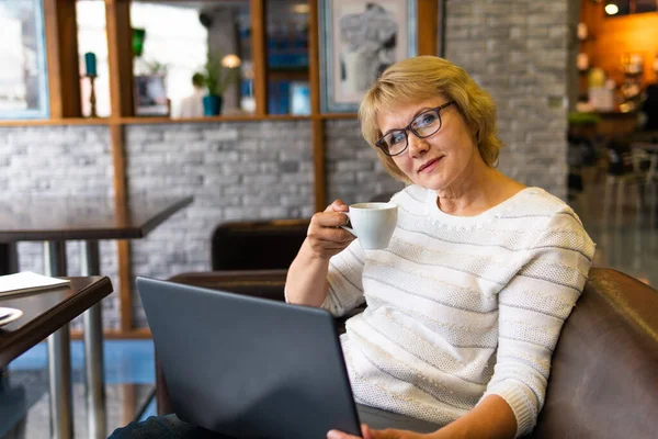 Una mujer sentada en una mesa con una taza de té y un portátil trabajando en un café . — Foto de Stock