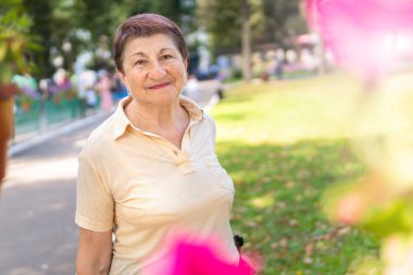 Yaşlı bir kadın, güneşli bir yaz gününde parkta sopayla kuzeye doğru yürüyor.