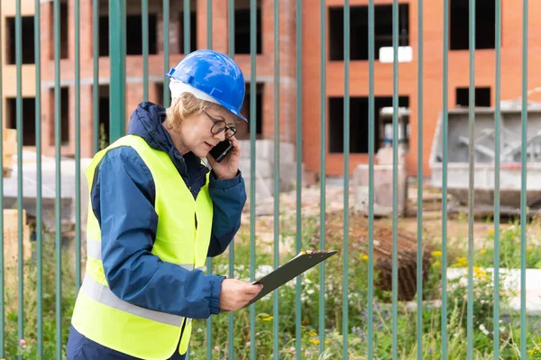 Builder woman at construction site in a green vest and a helmet with a tablet . A middle-aged woman with glasses inspects a new house.