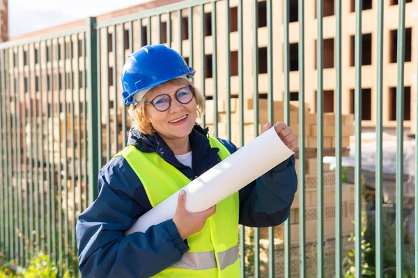 Builder woman at construction site in a green vest and a helmet with a project at home . A middle-aged woman with glasses inspects a new house.