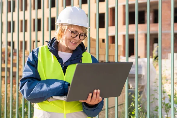A woman construction worker in a green vest and helmet with a laptop . A middle-aged woman with glasses inspects a new house.