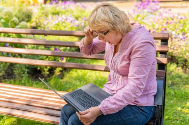 Parkta dizüstü bilgisayarda çalışan bir kadın. Orta yaşlı bir kadın haberleri izliyor. Doğada bir bankta oturuyor..