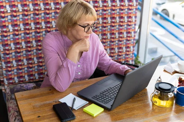 女性はカフェ オフィスのノートパソコンで働いています 中年の女性がお茶を飲んでニュースを見る 彼女はインターネットで話してる — ストック写真