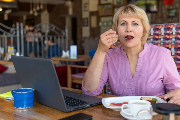 A woman works at a laptop in a cafe. A middle-aged woman drinks tea and watches the news. She\'s eating dessert.