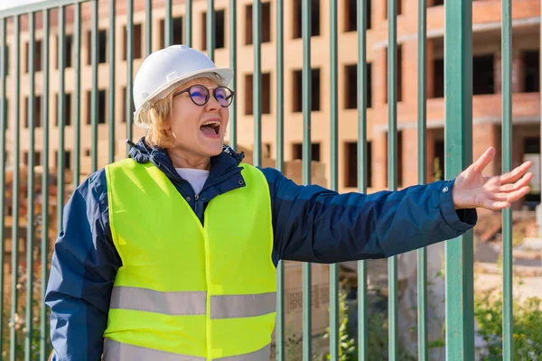 Female construction worker at a construction site in a green vest and helmet. A middle-aged woman with a folder inspects a new house. It forbids.