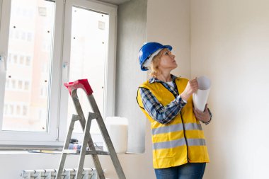 Kadın bir inşaat işçisi binanın tasarımını kontrol ediyor. Kasketli orta yaşlı bir kadın dairede tamirat yapıyor..