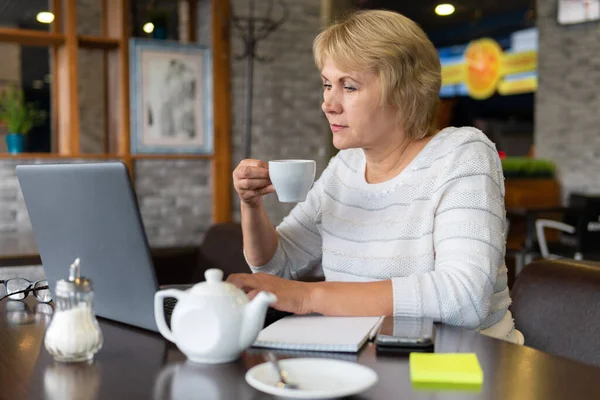 A woman works at a laptop in a cafe, office. A middle-aged woman drinks tea and watches the news. She talks over the Internet.
