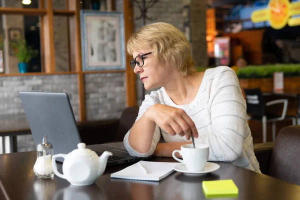女性はカフェ オフィスのノートパソコンで働いています 中年の女性がお茶を飲んでニュースを見る 彼女はインターネットで話してる — ストック写真