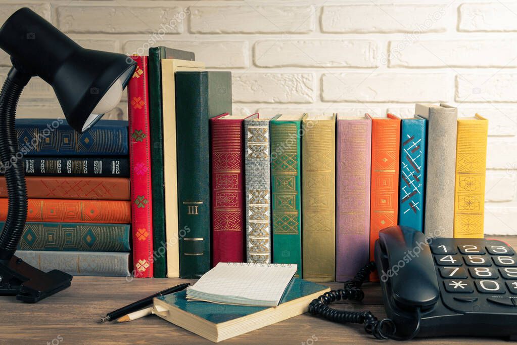 There are books of different colors and sizes on the bookshelf. On the table there is a phone, a Desk lamp and a notebook.