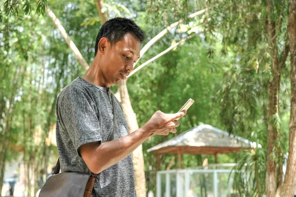 Молодий азіат дивиться на екран мобільного телефону. Привабливий молодий азіат дивиться на мобільний екран у парку. — стокове фото