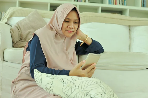 Wanita muslim tersenyum sambil membaca pesan di telepon pintar di ruang tamu — Stok Foto