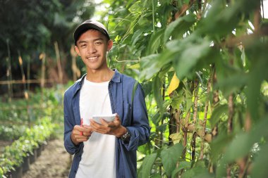 Mutlu gülümseyen Asyalı çiftçi erkek yeşil bahçede defterini tutuyor.