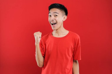 Genç Asyalı genç kazanan jest yapıyor, Happy özel bir hediye alıyor, kırmızı arka planda izole edilmiş.