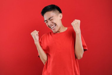 Genç Asyalı genç kazanan jest yapıyor, Happy özel bir hediye alıyor, kırmızı arka planda izole edilmiş.