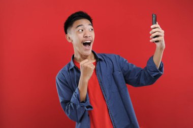 Akıllı telefon tutan Asyalı genç, elinde cep telefonu tutarak kazanıyor Happy, kırmızı arka planda izole edilmiş özel bir hediye alıyor.
