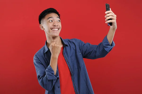 Akıllı telefon tutan Asyalı genç, elinde cep telefonu tutarak kazanıyor Happy, kırmızı arka planda izole edilmiş özel bir hediye alıyor.