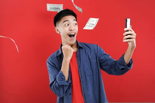Akıllı telefon tutan Asyalı genç, elinde cep telefonu tutarak kazanıyor Happy, kırmızı arka planda izole edilmiş özel bir hediye alıyor.