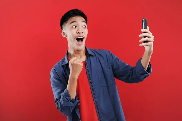 拿着智能手机的年轻亚洲少年拿着手机做着获胜的手势 快乐在网上得到特别的礼物 被红色背景隔离了 — 图库照片