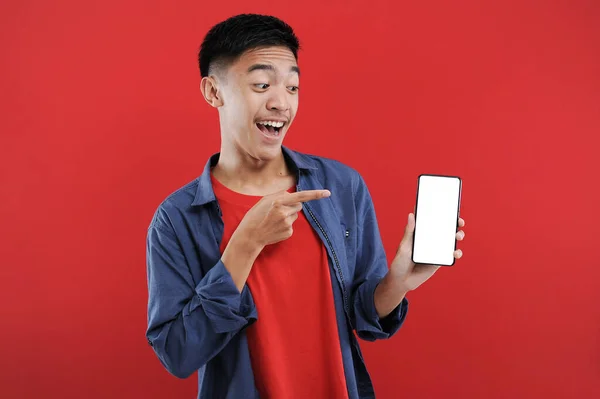 年轻的亚洲少年拿着带有空白屏幕的手机做着获奖手势 快乐地在网上买到特别的礼物 — 图库照片