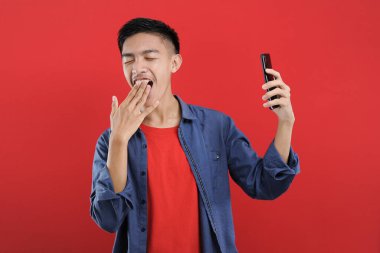 Uykulu poz veren Asyalı bir adam elinde akıllı telefon, kırmızı arka planda izole edilmiş.