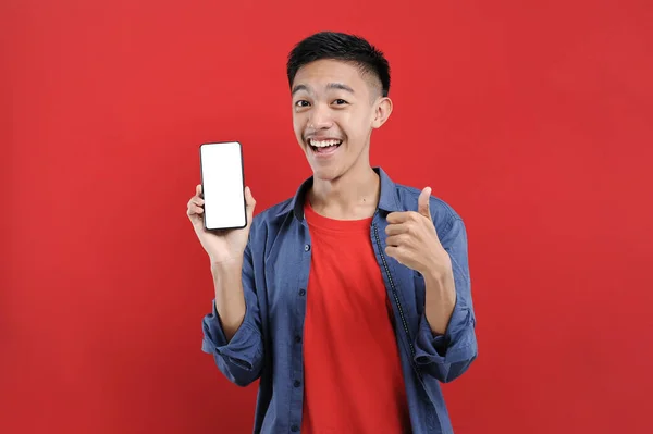 年轻的亚洲少年拿着手机做着获胜的手势 快乐在网上得到特别的礼物 被红色背景隔离 — 图库照片