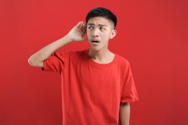 Kulağı havada Asyalı bir adam kırmızı arka planda izole edilmiş dedikoduları ve dedikoduları dinliyor.