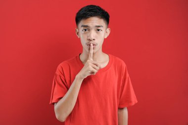 Tişörtünü giymiş, dudaklarına işaret parmağı tutarak sessiz kalmasını ve ses çıkarmamasını isteyen endişeli Asyalı genç bir adam. İnsan yüz ifadeleri.