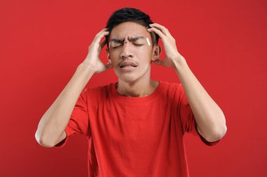 Baş ağrısı çeken genç Asyalı bir adam, başını tutarak acı çeken Asyalı bir gencin stüdyo portresi. kırmızı renkte
