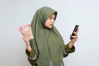 Genç Asyalı Müslüman Kadın, elinde cep telefonu ve para tutarak kazandığı jestleri mutlu ve heyecanlı bir şekilde ifade ediyor. Başarılı ve gri arka planda kutlama