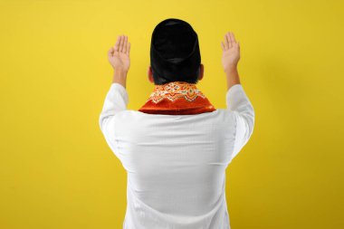 Genç Asyalı Müslüman adam elini kaldırıyor ve dua ediyor, geriye bakıyor, sarı arka planda izole edilmiş.