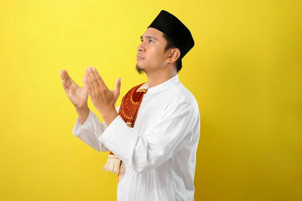 若いアジアのイスラム教徒の男性は 黄色の背景に隔離されたイスラム教徒のドレスで手を上げて離れて見て祈る — ストック写真