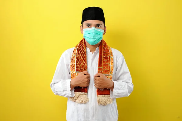 Korunan maske takan genç bir Asyalı Müslüman adam. Omzunda seccade var. Sarı arka planda izole edilmiş.