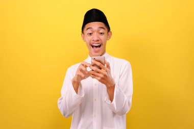 Mutlu Asyalı Müslüman adam, elinde cep telefonu olan Müslüman kıyafetlerle sarı arka planda izole edilmiş telefon ekranına bak.