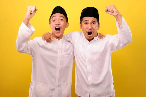 二つのハンサムな幸せなアジアのイスラム教徒の男性抱擁し 黄色の背景に隔離された勝利のジェスチャーを示しながら カメラを見て満足そうに顔 — ストック写真