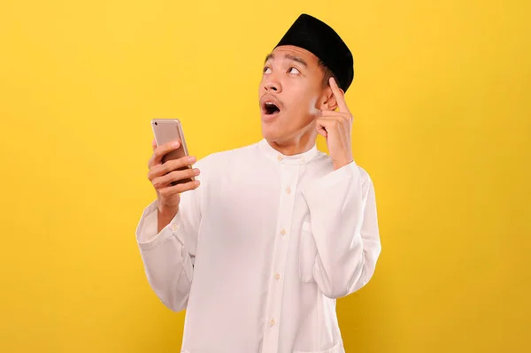 スマートフォンを持っている衝撃的な若いアジア系ムスリムの男のイメージと黄色の背景に隔離されたアイデアを考えます — ストック写真