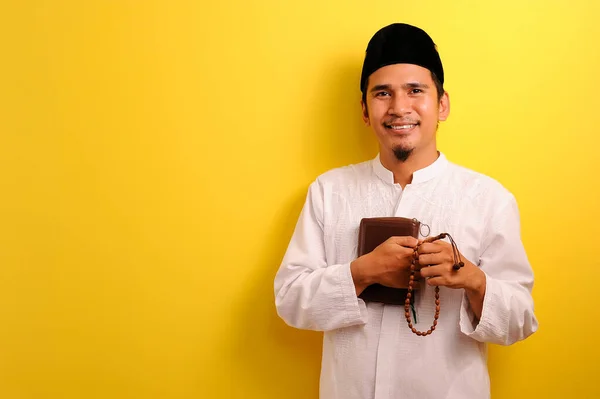 幸せな若いアジアのイスラム教徒の男性がクルアーンを保持し 黄色の背景に隔離されたコピースペースを持つビーズを祈る肖像画 — ストック写真