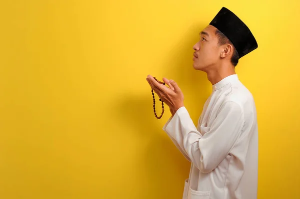 黄色の背景に隔離された祈りのビーズを保持して手を上げて祈る若いアジアのイスラム教徒の男性の別の角度 — ストック写真