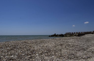 Rıhtım ve kayalar. Ahşap parmaklıklarla donatılmış bir rıhtım var. Mavi gökyüzü ve deniz