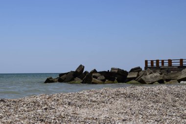 Denize giden köprü. Sudaki kayalar. Mavi deniz suyunda parla. Kumsalda kum ve deniz kabukları.