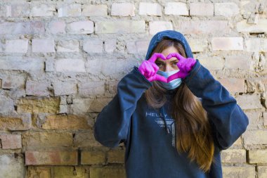 Mavi tıbbi maskeli kız terk edilmiş bir duvarın arkasında elleri pembe eldivenli aşk sembolü kalp işareti yapıyor. Coronavirus konsepti
