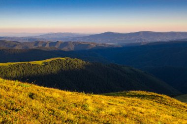 Bulutları aşan güneşin altında bir sıradağ. Güneş ışınları bulutları delip geçiyor ve eğimli dağlardaki kozalaklı yeşil bir ormanda parlıyor..