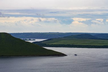 Gemi sarp kıyılar ve yeşil çimlerle kaplı pelerinler arasında dolambaçlı bir nehir yatağı boyunca ilerliyor. İlkbaharda nehrin üzerinde güzel bulutlar.