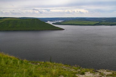 Nehrin manzarası, yeşil tepeler ve bulutlar. Sarmal bir nehir yatağı sarp kıyılardan ve yeşil çimlerle kaplı pelerinlerden akar. Nehrin üstündeki gökyüzünde yağmur bulutları.