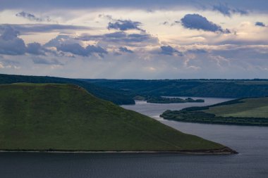 Nehrin manzarası, yeşil tepeler ve bulutlar. Sarmal bir nehir yatağı sarp kıyılardan ve yeşil çimlerle kaplı pelerinlerden akar. Nehrin üstündeki gökyüzünde yağmur bulutları.