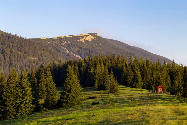 A small wooden chapel on the edge of a coniferous forest in an alpine meadow against the background of the sky and mountains at sunrise. A chapel in a forest in the mountains at dawn in the summer — Stock Photo, Image